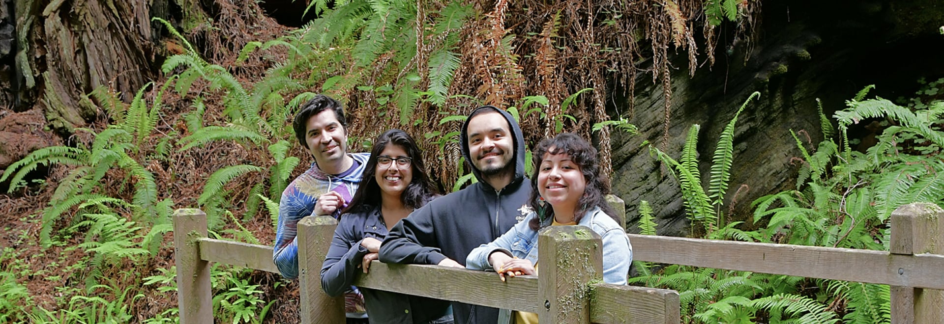 A cheerful family poses together leaning on a wooden fence amidst lush green ferns, with the majestic backdrop of Trillium Forest, enjoying their time in the serene natural setting.