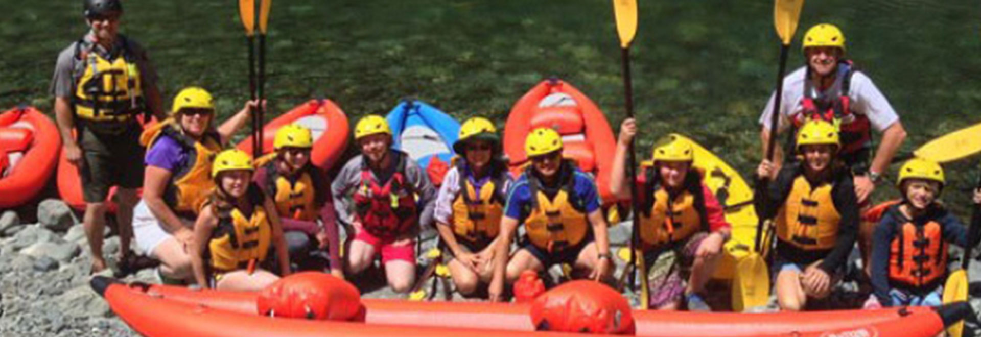A group of excited participants with paddles on a gravel river bank, ready for a Ranger-led kayak tour, geared up in life vests and helmets.
