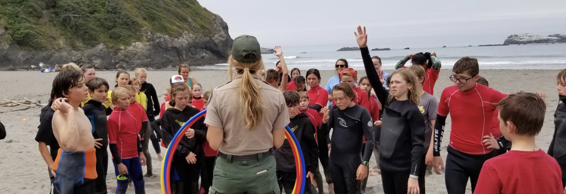 An attentive group of young students in wetsuits gather around a park ranger on a sandy beach, engaging eagerly in an educational outdoor activity with the ocean waves gently breaking in the background and a rocky island visible in the distance.