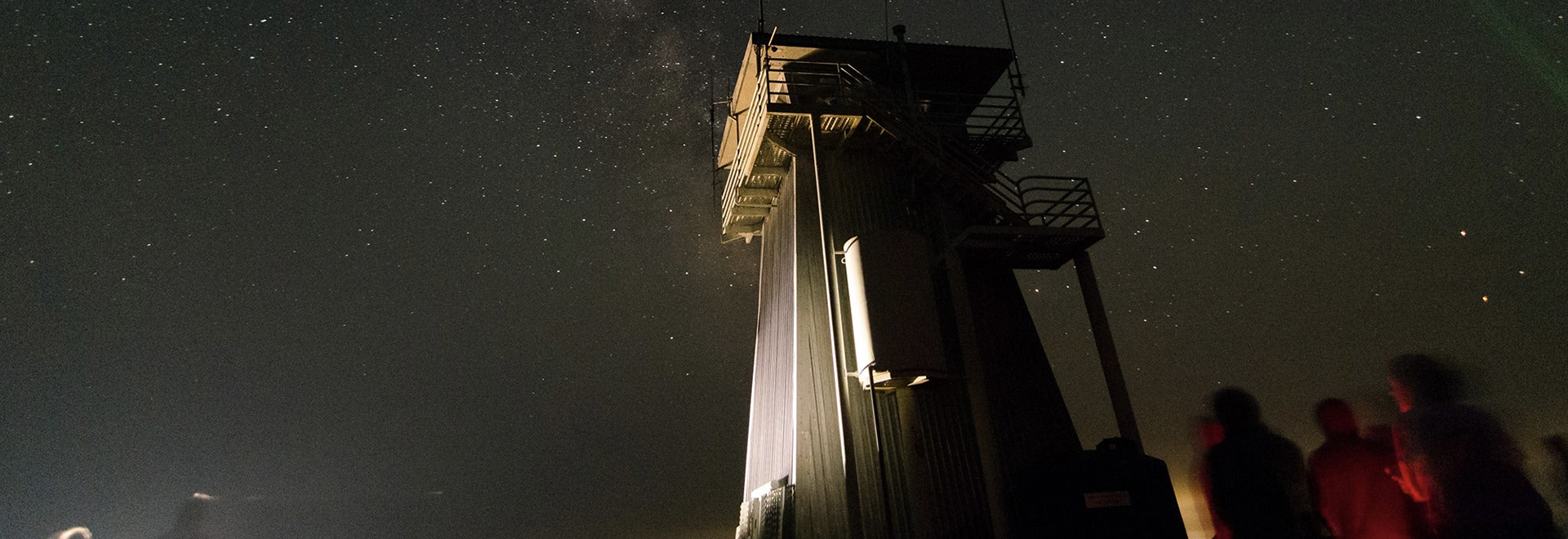 A group of people gather at night under a starry sky near a tall lookout tower, participating in a volunteer event at a park, marveling at the celestial wonders above.